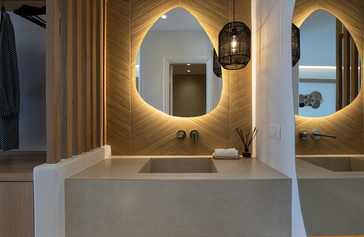 Bathroom area in hotel room in Mykonos Town at Damianos Hotel with backlit oval mirror, natural wood wall finish and modern sink design